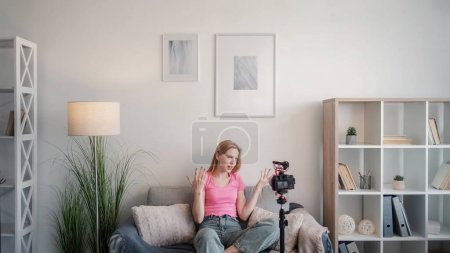 Photo for Video blog. Create content. Female expert. Young emotional influencer woman sitting on sofa talking to camera on tripod and gesticulating with hands in light interior at home free space. - Royalty Free Image