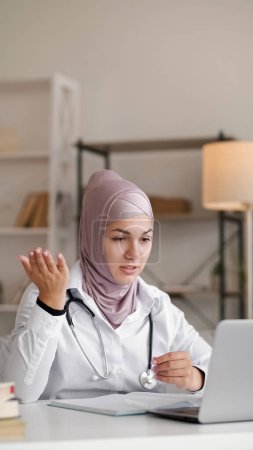 Foto de Médico en línea. Conexión de vídeo. Salud virtual. Mujer trabajadora médica en hijab con estetoscopio hablando en conferencia gesticulando en la mesa con portátil en habitación blanca. - Imagen libre de derechos