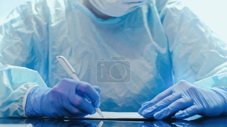 Photo for Disease history. Medical assessment. Coronavirus pandemic. Doctor in protective equipment writing report with hands in blue medical rubber gloves. - Royalty Free Image