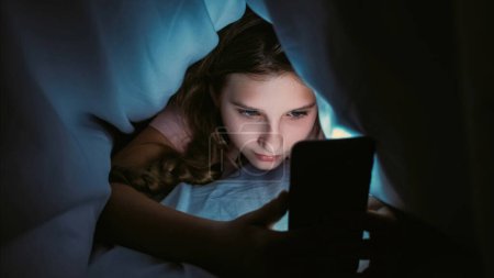 Photo for Screen time. Child insomnia. Late night chat. Internet addiction. Sleepless girl texting communicating on smartphone hiding under blanket in bed at home. - Royalty Free Image