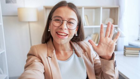 Photo for Video blog. Influencer streaming. Beautiful happy expressive young woman with big smile in joyful mood in glasses waving hand to camera for warm greeting in white room. - Royalty Free Image