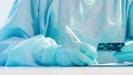 Photo for Drug prescription. Doctor medications. Healthcare paperwork. Nurse in blue protective equipment filling in form with pills blister in hand on white table. - Royalty Free Image