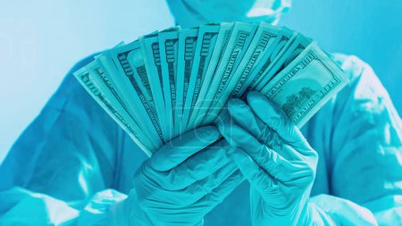 Healthcare costs. Medical expenses. Doctor salary. Surgeon hands in protective equipment holding US dollar cash money fan on blue gradient background.