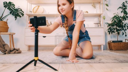 Photo for Kid vlog. Blogger home lifestyle. Create content. Happy smiling child girl adjusting phone camera on tripod recording video in light interior. - Royalty Free Image