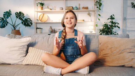 Photo for Kid blog. Influencer content. Online call. Enthusiastic girl recording speech for internet channel sitting with crossed legs on sofa in light room at home - Royalty Free Image
