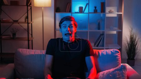 Photo for Grimacing man. Facial expression. Silly behavior. Foolish guy making nuts scare face sitting sofa in dark neon light home interior. - Royalty Free Image