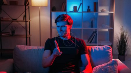 Photo for No gesture. Skeptic man. Facial expression. Disagreed smiling guy waving finger sitting sofa in dark neon light home interior. - Royalty Free Image