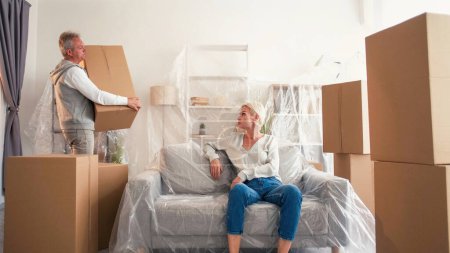 Foto de Mueva el apartamento. Desempacar pertenencias. Mujer sofá sentado en película de plástico cuando el marido toma cajas de artesanía propiedad en el interior de la habitación de luz. - Imagen libre de derechos