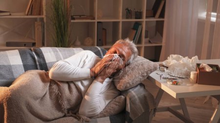 Foto de Goteo nasal. Gripe casera. El hombre que sufre estornudando síntomas de gripe dura acostado en el sofá bajo cuadros en el interior de la sala de sombra de luz. - Imagen libre de derechos