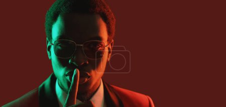 Photo for Man secret. Stop talking. Keep quiet. Neon light confident serious gentleman in suit glasses warning with hush gesture on dark red empty space background. - Royalty Free Image