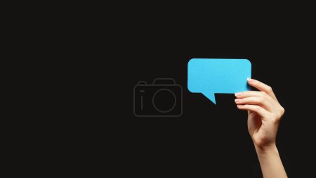 Photo for Dialogue icon. Internet chat. Network message. Female hand showing blue empty speech balloon isolated on dark black copy space background. - Royalty Free Image