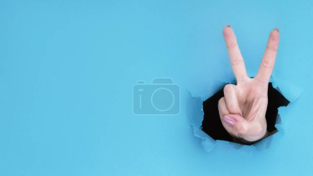Téléchargez les photos : Un geste de paix. Signe de victoire. Main féminine montrant numéro deux avec doigts à l'intérieur trou de papier percée sur fond bleu mur déchiré avec espace vide. - en image libre de droit