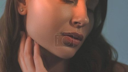 Photo for Lips makeup. Beauty trends. Plumping gloss. Balm shine. Unrecognizable woman with curvy puffy mouth in juicy lipstick with hand touching glowing radiant skin on grey background. - Royalty Free Image
