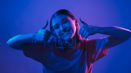Photo for Like gesture. Cool offer. Positive recommendation. Happy smiling woman in vibrant neon light showing thumbs up on blue background. - Royalty Free Image