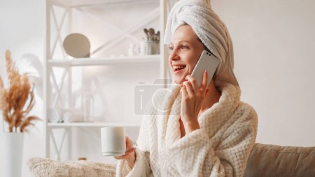 Foto de Mañana llamada. Comunicación móvil. Fin de semana de ocio en casa. Mujer sonriente alegre en toalla de albornoz con taza de café hablando en el teléfono en el interior de la luz con espacio libre. - Imagen libre de derechos
