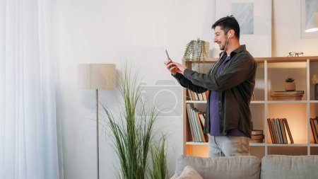 Photo for Phone video chat. Internet call. Home stream. Cheerful smiling man in wire earphones talking on mobile online web meeting in living room copy space. - Royalty Free Image