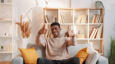 Photo for Approval sign. Apartment excitement. Recommend gesture. Happy smiling young man with thumbs up hands satisfied with flat interior on cozy sofa in living room at home. - Royalty Free Image