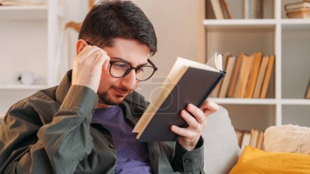 Photo for Book plot. Hard literature. Poor eyesight. Tensed frowning man in glasses struggling with complicated confusing paper novel on couch at home. - Royalty Free Image