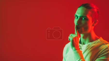Photo for Hush gesture. Man secret. Keep quiet. Green red color neon light smiling guy warning with shh finger at mouth on bright empty space background. - Royalty Free Image