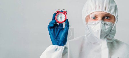Photo for Timely diagnosis. Virus specialist. Female woman doctor in medical protective equipment with alarm clock isolated on grey background empty space. - Royalty Free Image