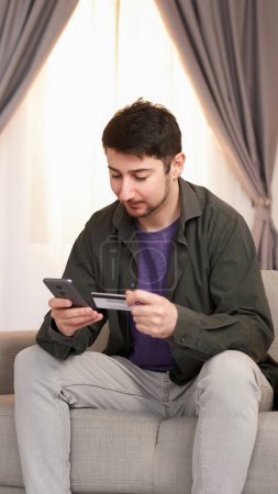 Photo for Money transfer. Online purchase. Internet shopping. Satisfied young man sitting with smartphone and bank credit card buying paying on sofa at home. - Royalty Free Image