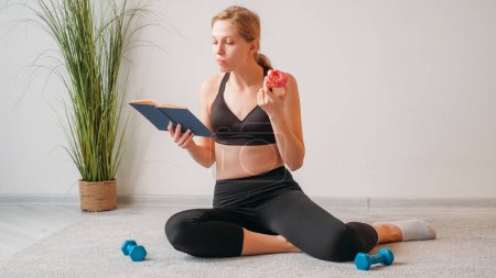 Photo for Fitness reading. Training break. Workout snack. Curious athletic woman with book eating sweet doughnut dessert on floor with dumbbells on light empty space. - Royalty Free Image
