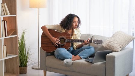 Foto de Práctica musical. Curso de guitarra. Estudiante. Mujer concentrada estudiando instrumento de cuerda en línea en el sofá sentado portátil en casa. - Imagen libre de derechos