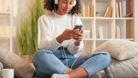 Photo for Music player. Home leisure. Internet browsing. Smiling woman in headphones scrolling smartphone at cozy sofa home living room. - Royalty Free Image