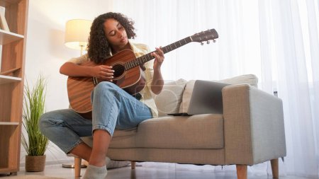 Photo for Song learning. Guitar playing. Music hobby. Focused woman musician practicing chords on acoustic instrument sitting couch with laptop home room. - Royalty Free Image