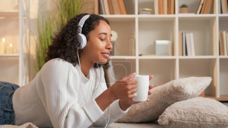 Photo for Cozy relax. Morning coffee. Hygge lifestyle. Cheerful dreamy woman with cup of drink in headset resting on floor at comfortable home living room. - Royalty Free Image