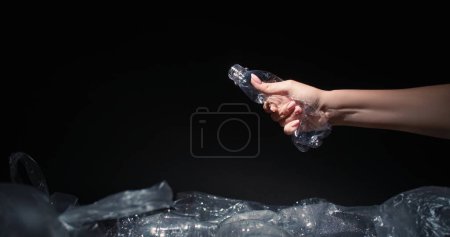 Téléchargez les photos : Réduction des déchets. Recyclage plastique. Femme volontaire main écrasant bouteille usagée jetant dans la poubelle avec des sacs de cellophane isolé sur fond noir espace vide. - en image libre de droit