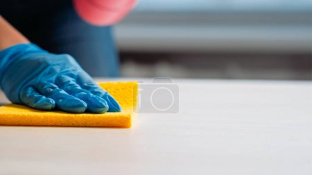 Photo for Cleaning company. Janitor services. Housekeeper hand in protective gloves washing light table furniture with surface wiping sponge carefully. - Royalty Free Image