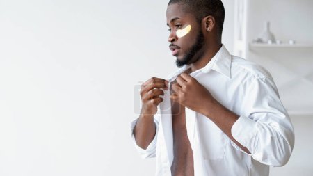 Photo for Male morning. Facial care. Relaxed confident handsome man with gold hydrogel eye face skin patch putting on white shirt at light modern interior home empty space. - Royalty Free Image