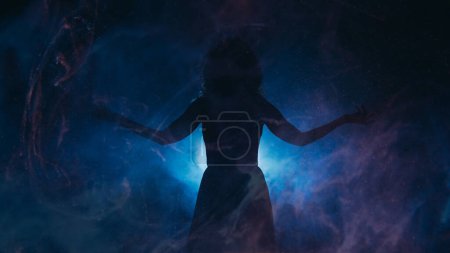Foto de Silueta de foco. Energía espiritual. Mujer con brazos extendidos en neblina sombra proyector azul brillante partículas niebla fondo oscuro espacio de copia. - Imagen libre de derechos