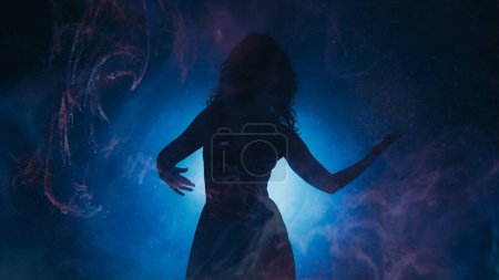Photo for Mysterious dance. Spiritual aura. Energetic woman silhouette moving in glowing swirls mist shadow in bright spot light on indigo background copy space. - Royalty Free Image