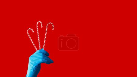 Photo for Candy canes. Pandemic holiday. Human hand in blue protective glove holding peppermint sweetmeats isolated on red copy space background. - Royalty Free Image