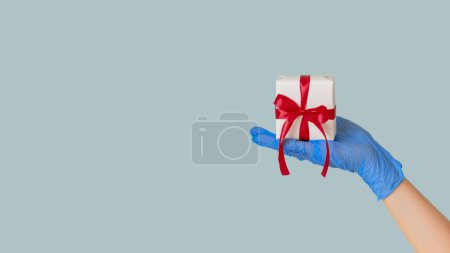 Téléchargez les photos : Livraison de cadeaux hygiène. Surprise du Nouvel An. Main féminine en gant de protection montrant enveloppé présent dans la boîte isolée sur fond d'espace de copie bleue. - en image libre de droit