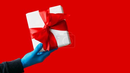 Photo for Festive gift. Present delivery. Unrecognizable human hand in protective gloves holding wrapped box isolated on red empty space background. - Royalty Free Image