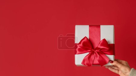 Foto de Sorpresa navideña. Un saludo romántico. Mano femenina mostrando regalo envuelto en caja blanca con lazo de cinta aislado sobre fondo de espacio de copia roja. - Imagen libre de derechos