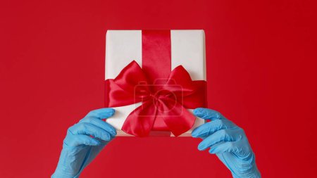 Photo for Christmas gift. Pandemic New Year. Female hands in blue medical gloves holding box with present isolated on red copy space background. - Royalty Free Image