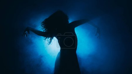 Foto de Coreografía contemporánea. Baile sensual. Mujer expresiva silueta corporal moviéndose en sombra de niebla oscura en el centro de atención en el espacio de copia de fondo índigo. - Imagen libre de derechos