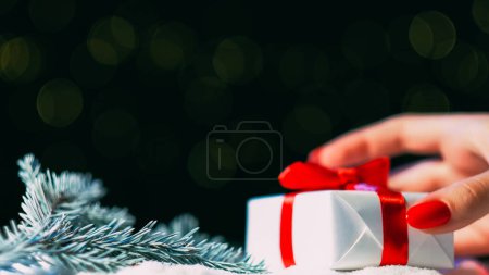 Foto de Regalo de Navidad. Sorpresa navideña. Felicitaciones de felicitación. Mujer mano poner presente caja cinta roja grabada en la rama de pino nevado decoración bokeh luces verdes espacio de copia de fondo. - Imagen libre de derechos