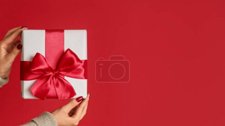 Téléchargez les photos : Surprise du Nouvel An. Cadeau de Noël. Main féminine montrant blanc enveloppé présent dans la boîte avec ruban arc isolé sur fond vide rouge. - en image libre de droit