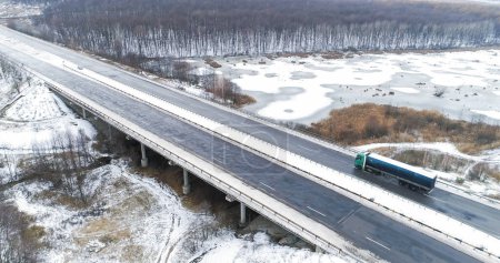 Photo for Aerial winter traffic. Drone countryside road. Snowy bridge trough frozen lake asphalted icy danger driveway in cold cloudy day. - Royalty Free Image