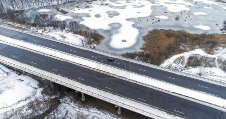 Photo for Aerial snowy bridge. Winter road. White frozen lakes and asphalted track traffic icy river water in cloudy morning cold season. - Royalty Free Image