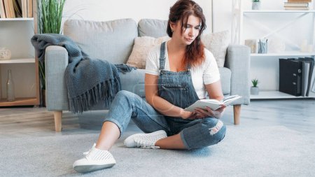 Photo for Home reading. Dreamful woman. Enjoying literature. Smiling middle-aged lady sitting floor with interesting book in light room interior. - Royalty Free Image