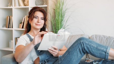 Photo for Home reading. Happy adult woman. Enjoying life. Smiling middle-aged lady laying sofa with interesting book in light room interior. - Royalty Free Image