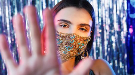 Photo for Covid party. Pandemic jewelry. Tender mysterious female model with night makeup in matching gold chain face mask outstretching hand on blue shiny tinsel. - Royalty Free Image