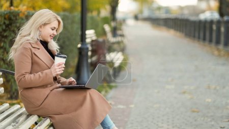 Photo for Freelance work. Happy woman. Virtual connection. Smiling lady sitting bench in park alley typing laptop holding cup of coffee copy space. - Royalty Free Image