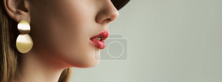 Téléchargez les photos : Maquillage des lèvres. Tendance cosmétique. Jeune belle femme méconnaissable en rouge à lèvres juteux parfait visage peau boucle d'oreille dorée sur fond gris espace vide. - en image libre de droit
